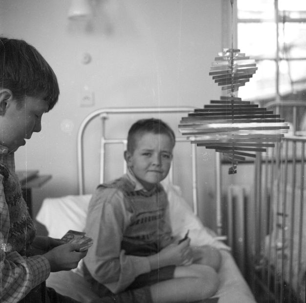 Black and white photograph. Two children seated on a bed in a hospital ward holding playing cards, one looking out at us. Next to them is a metal spiral-shaped mobile sculpture that makes two revolutions to form a shape that's narrow at the top and wide at the bottom. '