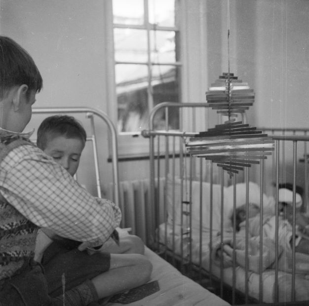 Black and white photograph. Two young children seated on a hospital bed playing cards next to a spinning metal mobile sculpture. Another child is asleep in a crib next to them.