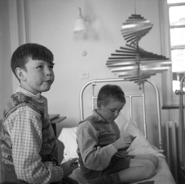 Black and white photograph. Two young children playing cards in a hospital bed, as one looks up with an interested expression at a metal mobile sculpture that spins and catches the light from a nearby window.