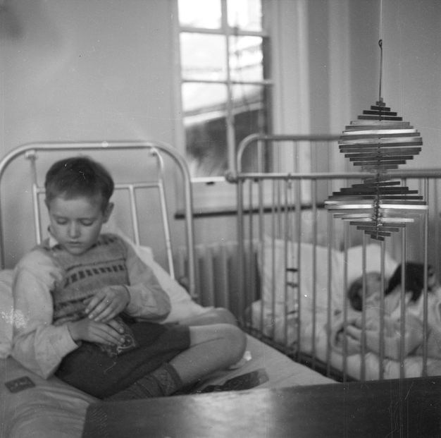 Black and white photograph. Young child seated in a hospital bed reaches over to pick up another playing card to add to their hand. A corkscrew-shaped metal mobile hangs next to the bed at just about eye-level with the child.