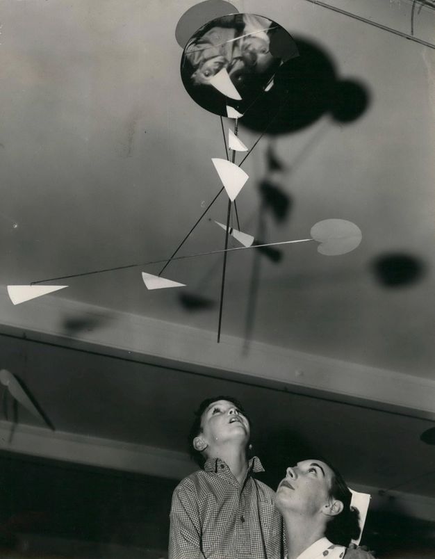 Black and white photograph. Nurse holding a child both look up at a mobile sculpture hanging from the ceiling. The sculpture consists of interlocking rods extending down and out in all directions with a few geometric shapes placed throughout the sculpture.