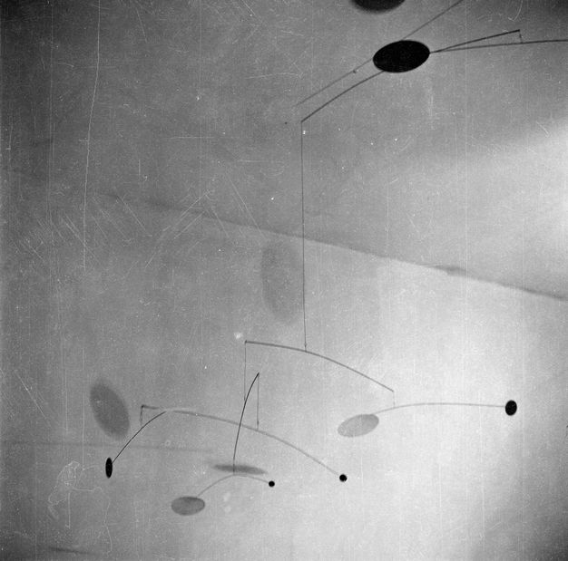 Metal sculpture. Black and white photograph looking up at a multi-tiered mobile sculpture hanging from the ceiling. Each tier consists of brackets extending horizontally out so far that the rods seem to bend with the weight of the geometric shapes dangling at each end.