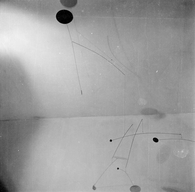 Metal sculpture. Black and white photograph looking at a partial view of a multi-tiered mobile sculpture, with brackets extending horizontally out, some that appear to bend more in one direction, perhaps with the unbalanced weight of geometric shapes at each end.