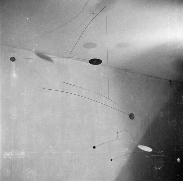 Metal sculpture. Black and white photograph looking up at a mobile sculpture hanging from the ceiling with multiple tiers of narrow curved metal rods and cables that offer a delicate and bouncy three-dimensional shape.
