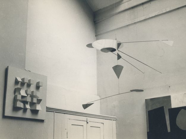 Exhibition. Abstract metal mobile sculpture with geometric shapes hangs from the ceiling next to a square-shaped panel fixed to the wall that contains a loose four by four grid of square and triangular blocks.