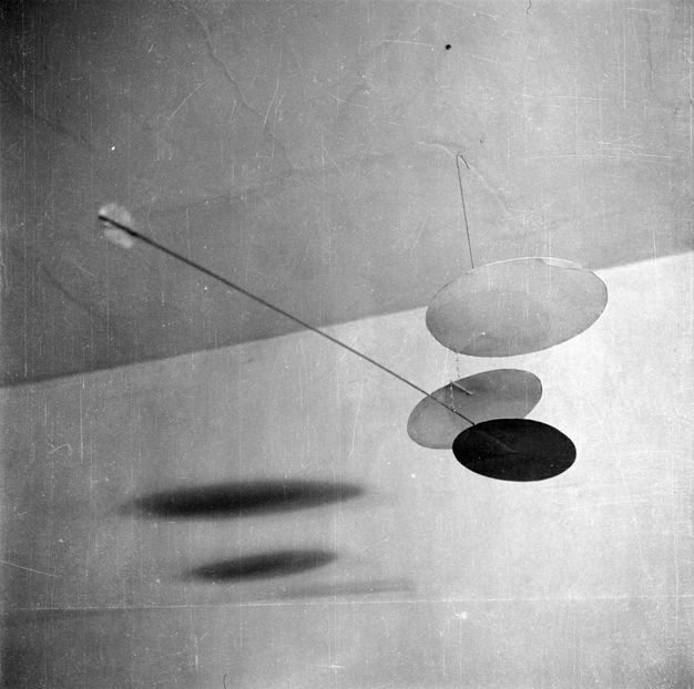 Mobile sculpture. Black and white photograph. A vertical rod hangs down from the ceiling connecting to a cluster of differently-sized circular shapes. A relatively longer rod extends diagonally up at an angle from the cluster to an individual circle at its endpoint.