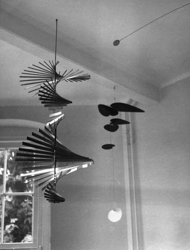 Installation. Black and white photograph of several mobile sculptures; one in the shape of a corkscrew that expands and contracts across four revolutions, the other a cluster of flat differently-sized geometric shapes extending out from a single vertical rod.