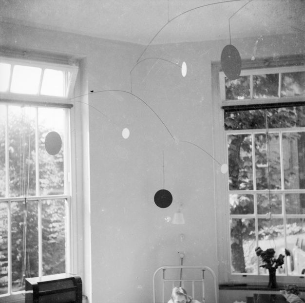 Metal sculpture. Black and white photograph of a delicate multi-tiered mobile sculpture that nearly fills the corner of a bright hospital ward. The mobile has long thin metal rods that extend horizontally out to circular shapes that reflect the light from a nearby window.