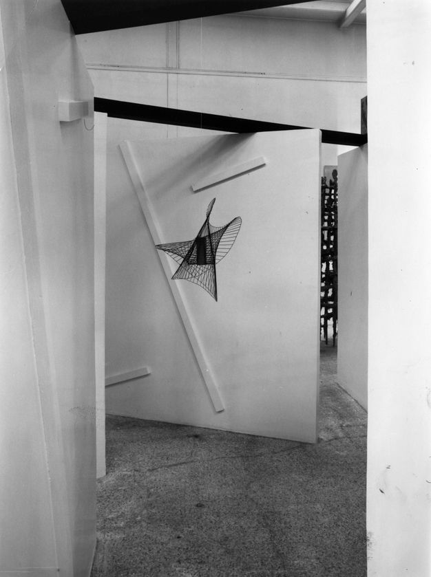 Exhibition. Black and white photograph of a view down an angled corridor towards a metal mobile sculpture that forms an abstract three-dimensional star shape with five curved points.