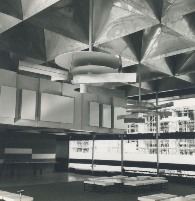 Architecture. Black and white photograph of an interior building filled with abstract designs, from the reflective three-dimensional grid ceiling, to the layered rectangular wall panels, and hanging mobile sculptures of circular plates and rectangular beams.