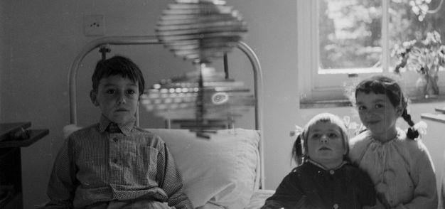 Black and white photograph. Three children in a hospital ward look towards us at a corkscrew-shaped metal mobile sculpture that dangles in the foreground. Two children are looking with dazzled expressions towards the mobile and the third is smiling sweetly at us.