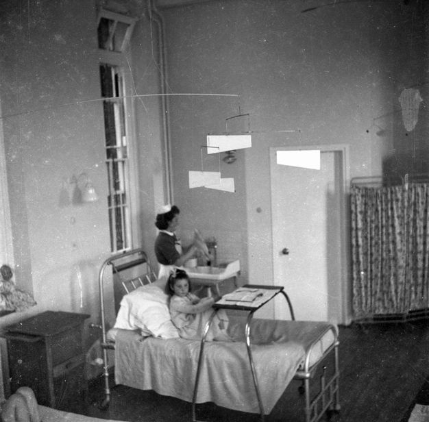 Black and white photograph. Child seated up in bed in a hospital ward with a tray table set up in front of them as a nurse works at a sink station beside them.