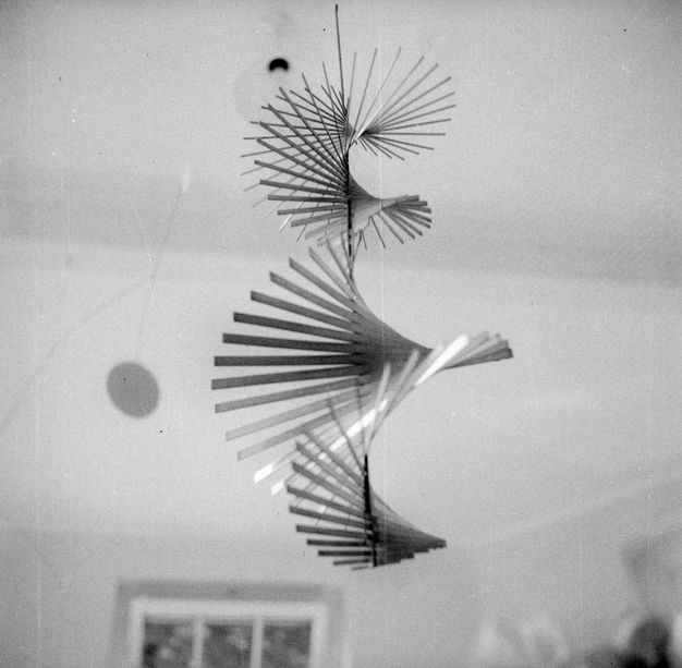Metal sculpture. Black and white photograph looking up the length of a spiral-shaped mobile sculpture hanging from the ceiling. From this angle we can see that the spiral shape makes three complete revolutions.