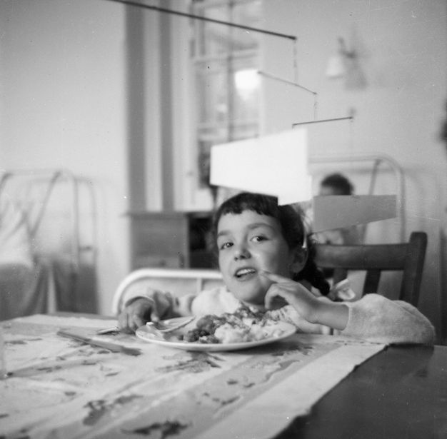 Black and white photograph. Young child smiles playfully across a relatively high dining table. There is a plate of food in front of them with a mobile hanging down from the ceiling just above their head.