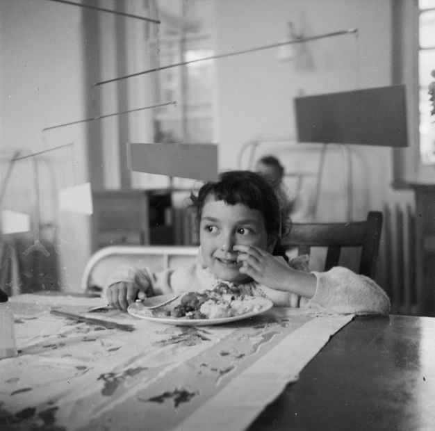 Black and white photograph. Child seated at a table in front of a plate of food with a mobile sculpture above that extends out in tiered brackets, each with a dangling trapezoidal shape.