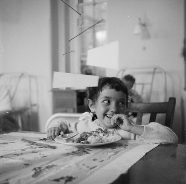 Black and white photograph. Child at a dining table looks away with delight as a multi-tiered mobile sculpture rotates above their head, each tier articulating to form a delicate three-dimensional shape.