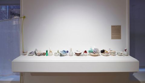 A display featuring small ceramic bowls, each filled with various found objects such as paper, fabric, and other mixed media materials, arranged on a white floating shelf.