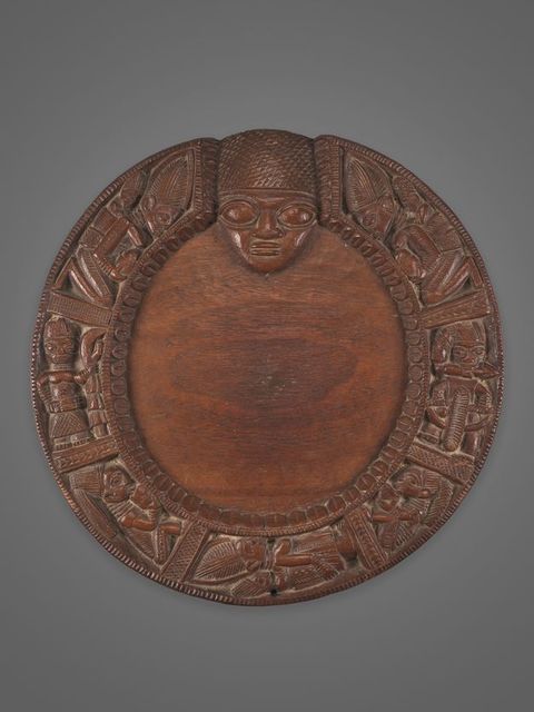 A circular wooden plaque with elaborate carvings of human and animal figures around the border, featuring a prominent human face at the top centre.