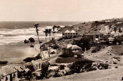 A black-and-white photograph of a coastal village with wooden houses, palm trees, a rocky shoreline, and waves breaking on the beach.