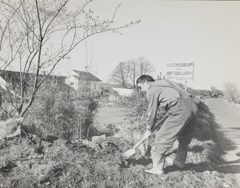 A black-and-white photograph of a man in overalls digging in an outdoor area. A sign in the background reads 'LYC Museum and Art Gallery'.