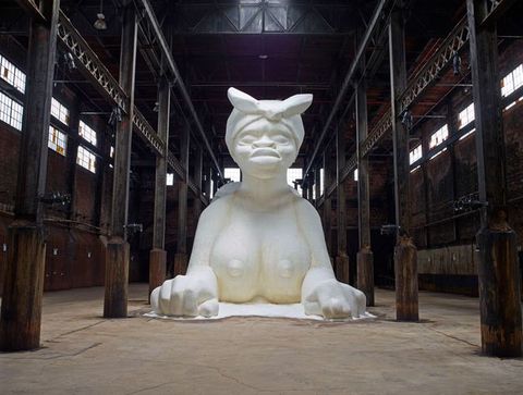 A monumental white sculpture of a Sphynx-like creature, made from sugar, inside an old industrial warehouse.