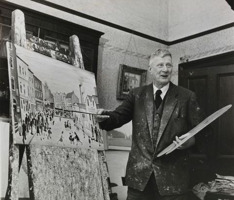 A black-and-white photograph of an older artist in a suit, holding a paintbrush and standing in front of an easel displaying a cityscape painting.