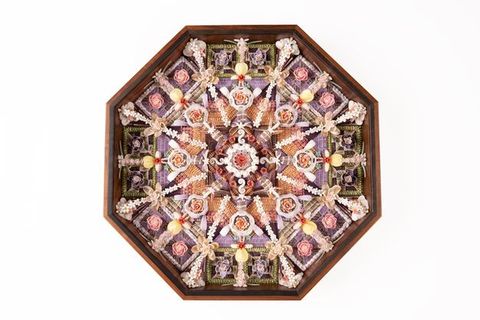 A symmetrical shellwork design in an octagonal wooden frame, featuring flowers, beads, and woven elements in shades of pink, purple, and beige.