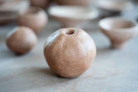 A close-up of a round, textured ceramic object with a small hole at the top, resembling a hollow clay sphere. Blurred in the background are more handmade ceramic pieces.