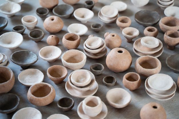 A collection of small handmade ceramic bowls and vessels in earthy tones, displayed on a wooden surface. The pieces vary in size and shape, with some stacked and others scattered.