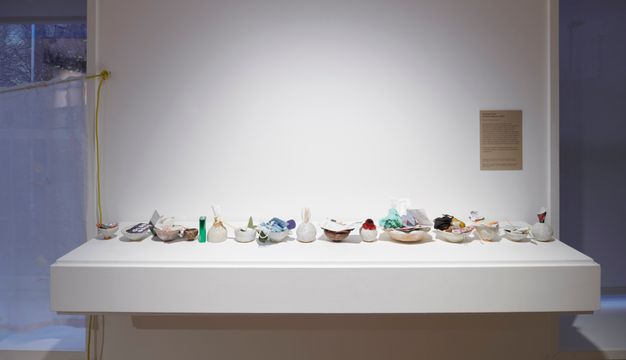 A display featuring small ceramic bowls, each filled with various found objects such as paper, fabric, and other mixed media materials, arranged on a white floating shelf.