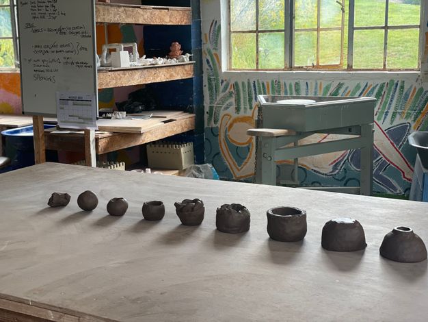 A row of small, hand-formed clay objects at various stages of shaping, placed on a wooden table in a pottery studio. The background features a colourful mural, a pottery wheel, and wooden shelves filled with ceramic pieces and tools. A whiteboard with handwritten notes and a calendar is also visible in the workspace.