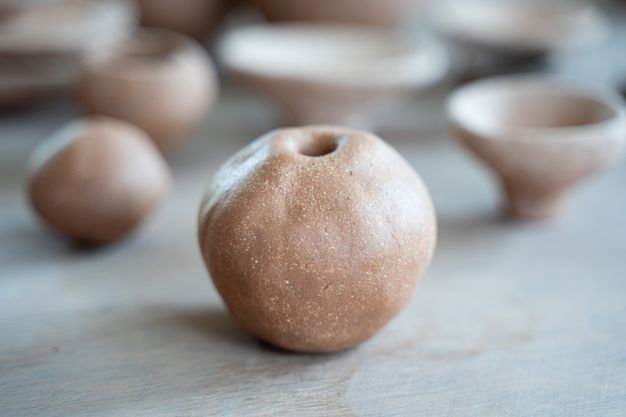 A close-up of a round, textured ceramic object with a small hole at the top, resembling a hollow clay sphere. Blurred in the background are more handmade ceramic pieces.