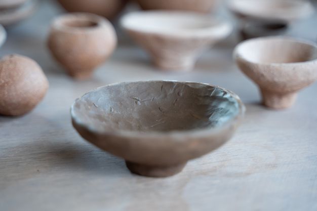 A small, brown, and rough-textured ceramic bowl with an uneven rim, sitting on a short pedestal base. Several other ceramic vessels are visible and blurred in the background.
