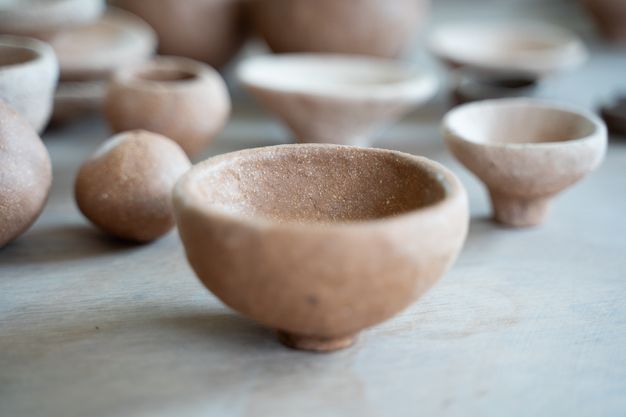 A small, light brown and rough-textured ceramic bowl, sitting on a short pedestal base. Several other ceramic vessels are visible and blurred in the background.