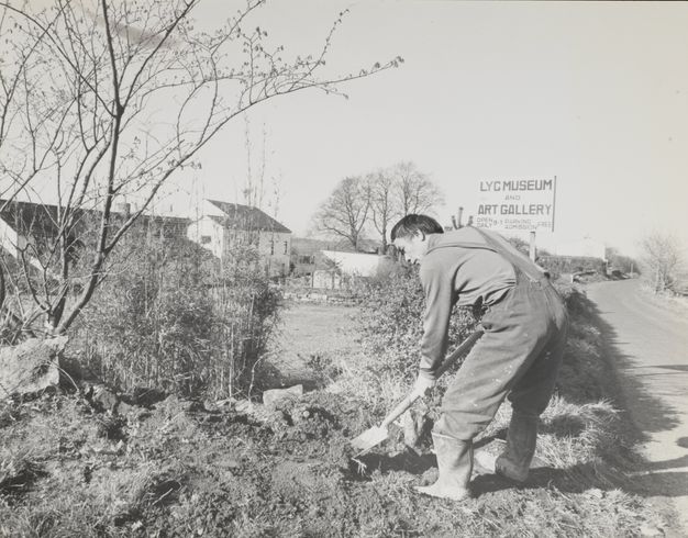 A black-and-white photograph of a man in overalls digging in an outdoor area. A sign in the background reads 'LYC Museum and Art Gallery'.
