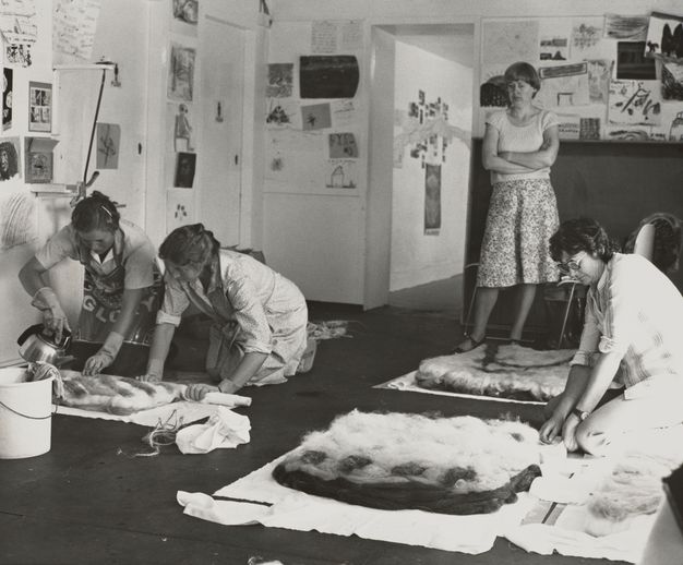 A black-and-white photograph of four women participating in an indoor creative workshop, working on felt pieces. The walls are covered in drawings and notes.