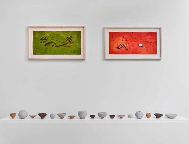 A gallery display featuring a row of small ceramic vessels placed on a white shelf, beneath two framed abstract paintings---one in green and the other in red.