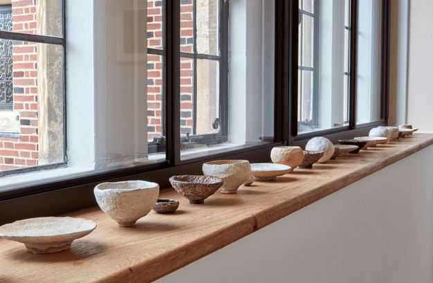 A row of ceramic bowls displayed along a wooden windowsill in a well-lit gallery space. The bowls vary in size, colour, and texture.