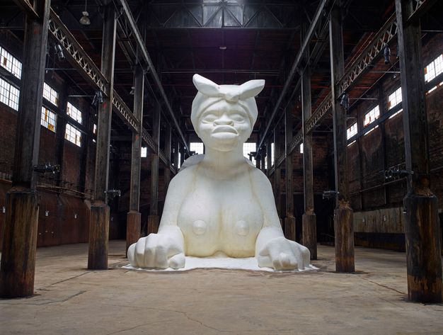 A monumental white sculpture of a Sphynx-like creature, made from sugar, inside an old industrial warehouse.