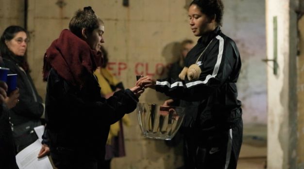 A photograph of a person wearing a black tracksuit and a woven wicker breastplate hands a small vessel to another person wrapped in a scarf, while others watch in the background.