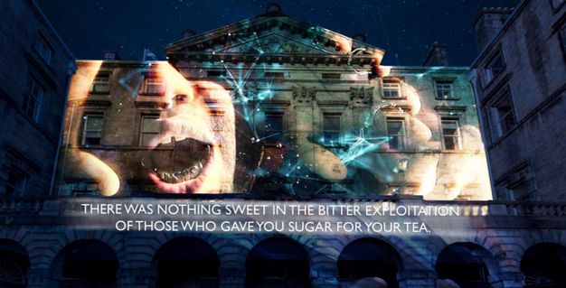 A nighttime projection on the façade of a grand historic building, displaying abstract imagery of faces, hands, and a cosmic web-like structure. The projection includes a bold message: 'THERE WAS NOTHING SWEET IN THE BITTER EXPLOITATION OF THOSE WHO GAVE YOU SUGAR FOR YOUR TEA'.