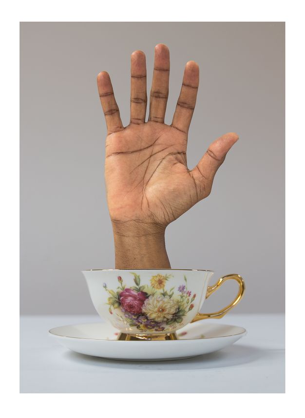 A delicate floral-patterned teacup with a human hand emerging from it, upright with fingers spread. The teacup sits on a matching saucer against a plain background.