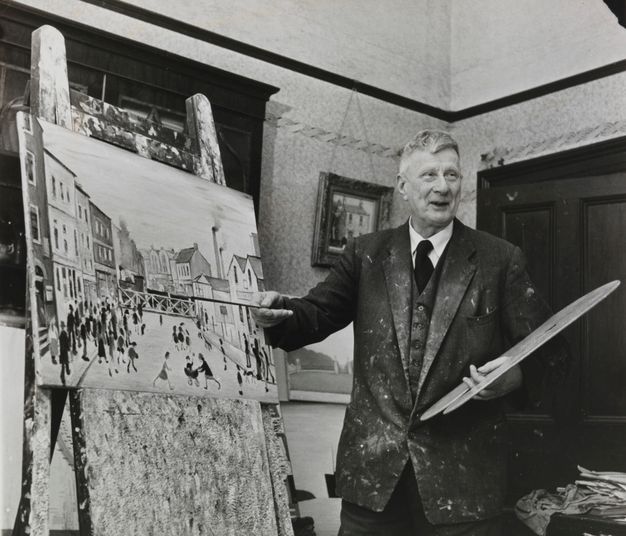 A black-and-white photograph of an older artist in a suit, holding a paintbrush and standing in front of an easel displaying a cityscape painting.
