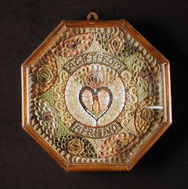 An octagonal sailor’s valentine encased in glass, featuring an intricate shell mosaic with a central heart motif surrounded by floral patterns in shades of white, pink, and brown.