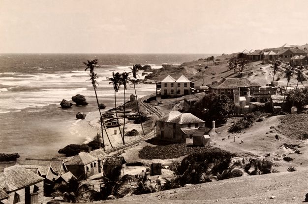 A black-and-white photograph of a coastal village with wooden houses, palm trees, a rocky shoreline, and waves breaking on the beach.