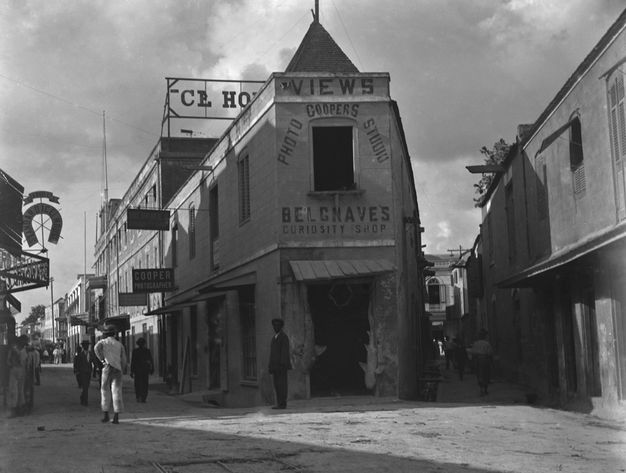A black-and-white street photograph showing a triangular-shaped building with signs reading 'Coopers Photo Studio' and 'Belgnave's Curiosity Shop,' surrounded by colonial-style buildings.