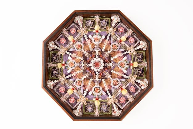 A symmetrical shellwork design in an octagonal wooden frame, featuring flowers, beads, and woven elements in shades of pink, purple, and beige.