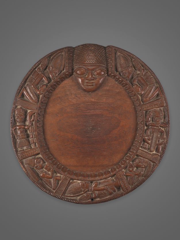 A circular wooden plaque with elaborate carvings of human and animal figures around the border, featuring a prominent human face at the top centre.