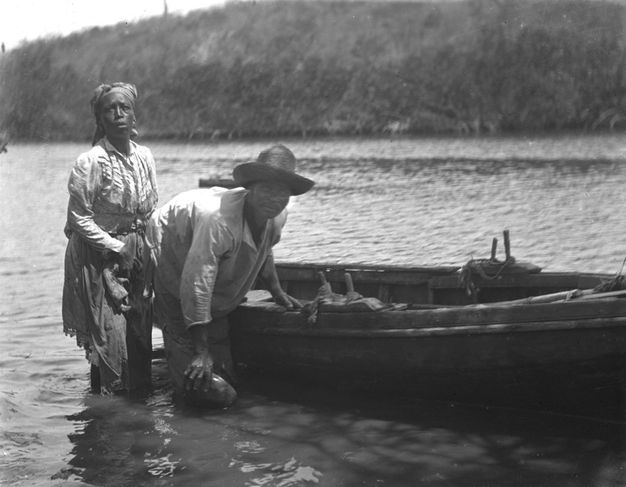 A black-and-white photograph of two people, a woman and a man, standing by a wooden boat in shallow water, wearing simple clothing and hats.