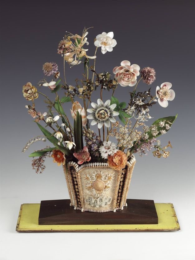 A decorative floral arrangement made entirely from shells, featuring sculpted flowers, leaves, and stems in a container also embellished with shells.